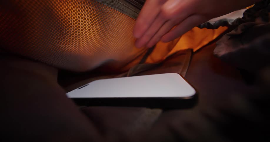 Hand reaches for whitescreen smartphone inside dark space lit by orange glow from meshlike surface above