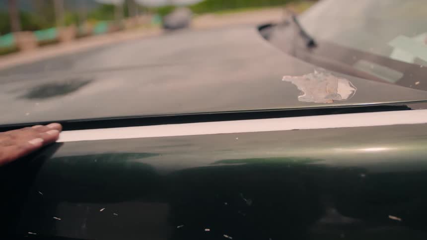 This is a closeup shot focused on the hood of a darkcolored car likely parked outside showing a detail of the vehicles condition a hand briefly rests on the edge of the hood drawing attention to a prominent area of peeling paint or clear coat near