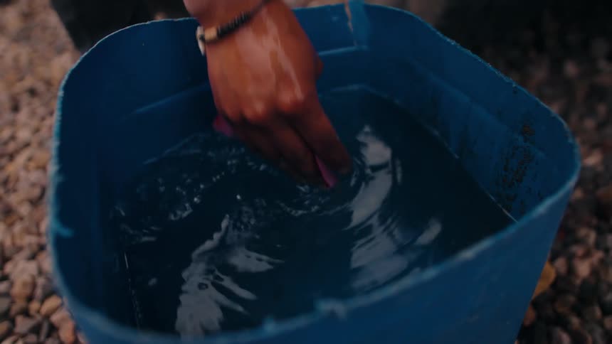 A hand wearing a beaded bracelet reaches into a large blue plastic barrel filled with water stirs the water with a vigorous movement and then quickly pulls the hand out of the barrel