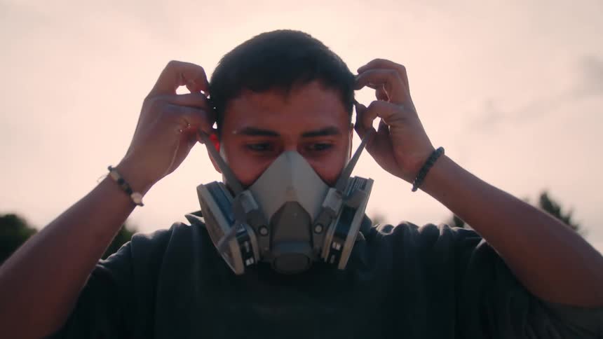 The video is a closeup lowangle shot of a person putting on a respirator mask with dual filter cartridges against a bright hazy sky background they are seen carefully adjusting the straps over their head and face indicating preparation for a task t