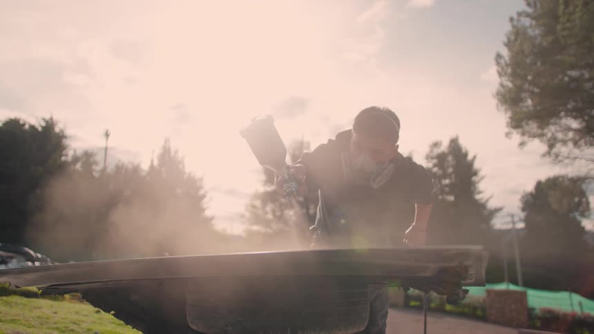 Person performs outdoor task with spray gun over panel supported by tire surrounded by dust mist under warm evening light