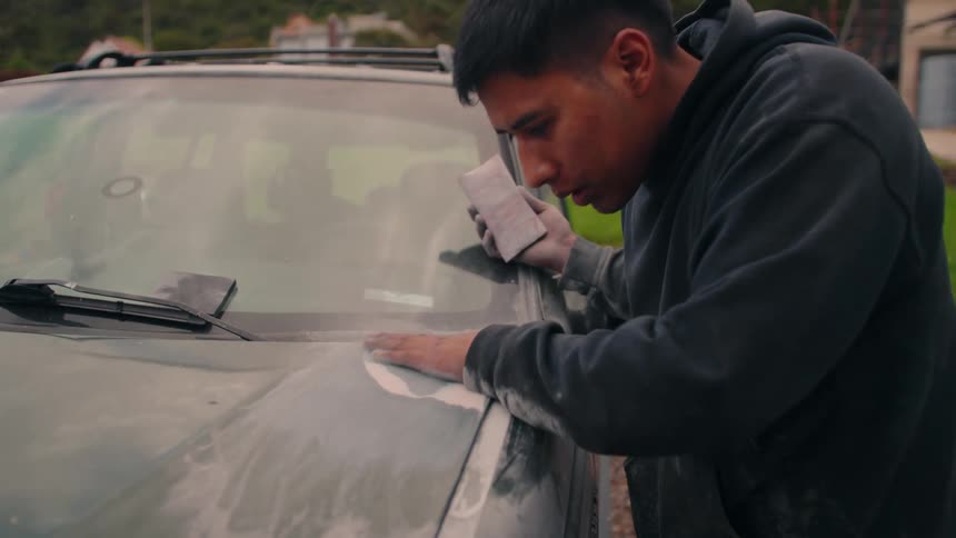 Person in dark hoodie sands car hood with primer patches in outdoor area with greenery in the background