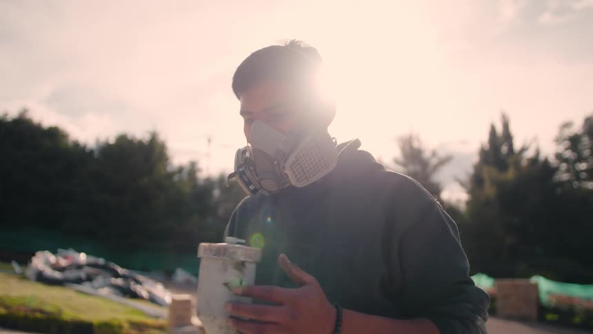 Man wearing respirator holds paint container during outdoor task under clear sky and trees in the background