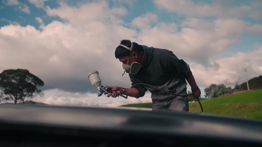 A man stands outdoors at midday wearing a protective mask as he carefully spray paints a metal piece focused on achieving a smooth even coat
