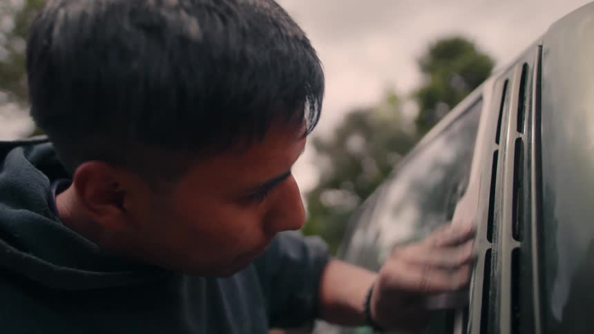 Man wipes car windshield with cloth in outdoor setting under cloudy sky and trees in the background