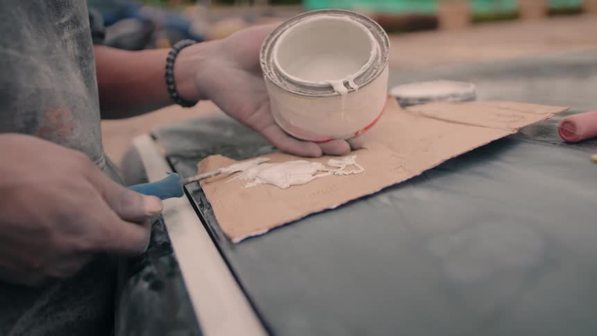The image is a closeup of a persons hands performing a task where one hand holds a small can of thick white paint or puttythe other hand uses a tool with a blue handle to scoop the substance out of the can and apply it to a piece of cardboard or su