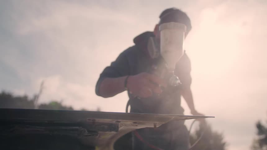 A person wearing a respirator holds a spray gun sprays a paint mist onto a dark object and works in bright outdoor lighting