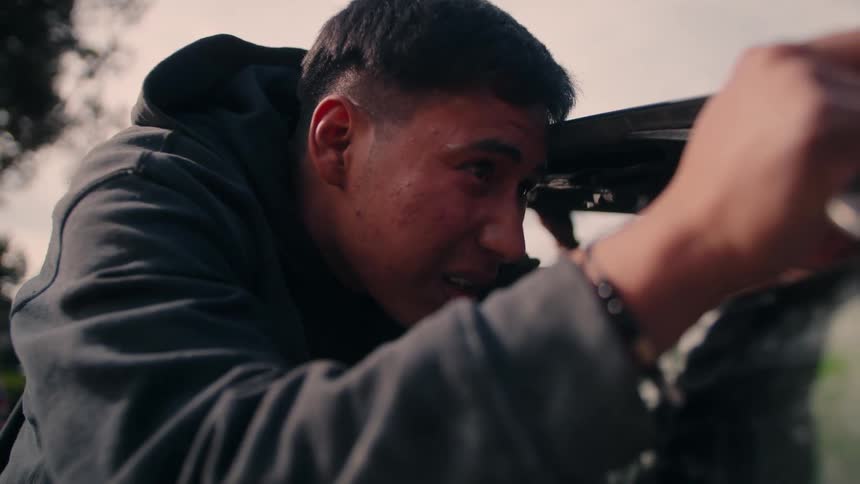 Man in dark hoodie works intently outdoors holding a tool while closely inspecting a part