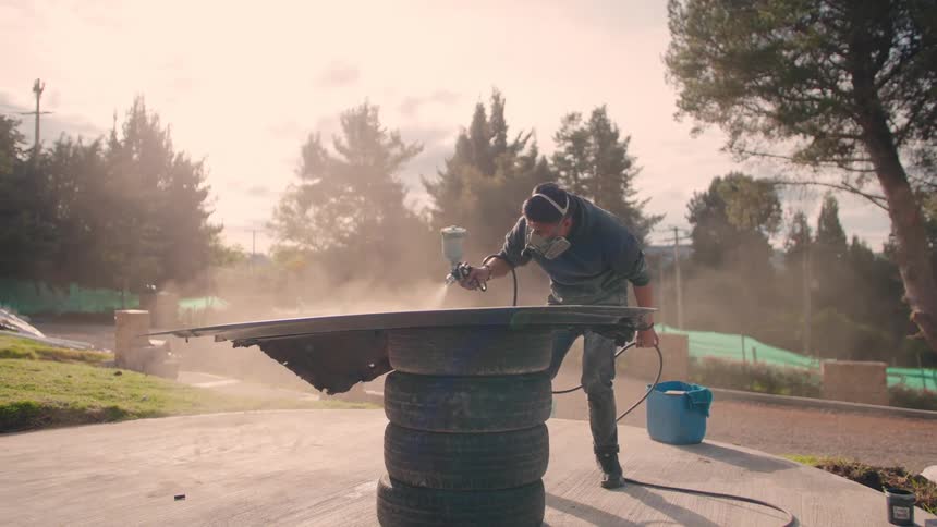 The video shows a person wearing a respirator mask and dark clothes standing outdoors and using a spray gun to paint a large dark object likely a car part resting on a stack of tires the scene is bright and dusty with trees and greenery visible in t