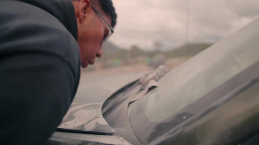 The video shows a closeup of a person seen in profile wearing a dark hoodie and glasses applying a layer of film or wrap to the exterior of a vehicle the person leans in to air onto the film likely to help flatten it or remove bubbles during the