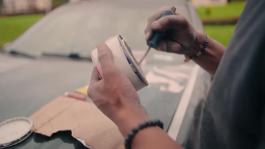 Man opens white container with blue rim using screwdriver on car hood with tools nearby