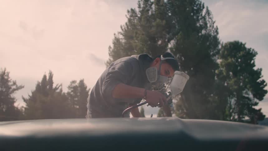Person wearing protective mask applies paint with spray gun on surface outdoors under clear sky and trees in the background