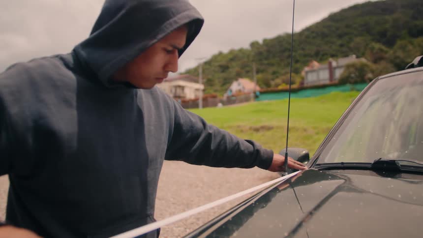 Man applies white tape on dark vehicle hood parked on gravel area with hills and cloudy sky in the background