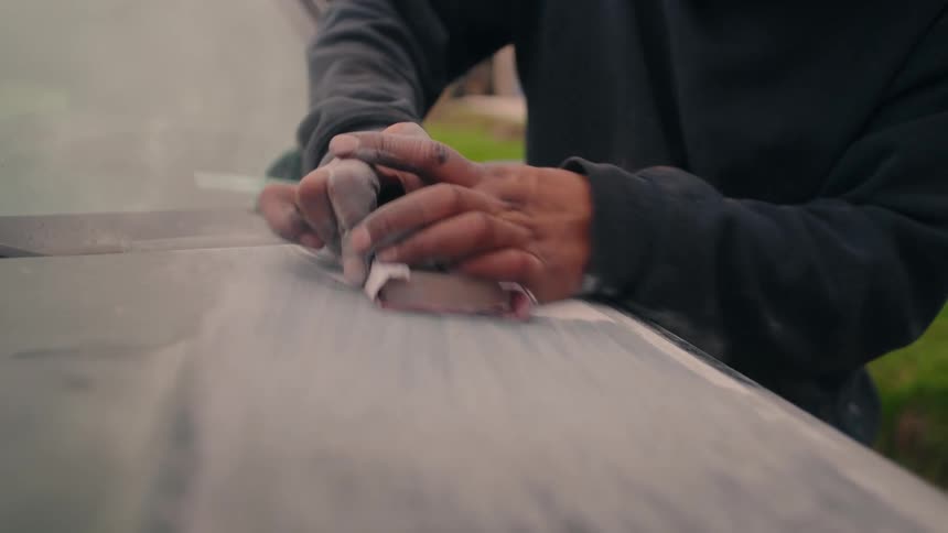 This is a closeup shot focusing on the hands of a person who is sanding a large smooth gray surface likely part of a vehicle or fiberglass object the person is using a small piece of sandpaper to vigorously smooth the primer or putty generating dust