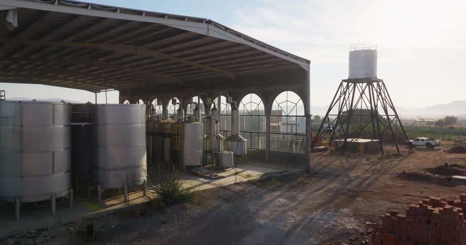 Cylindrical metal tanks occupy covered industrial tequila distillery facility beside elevated water tower on land overlooking distant hills