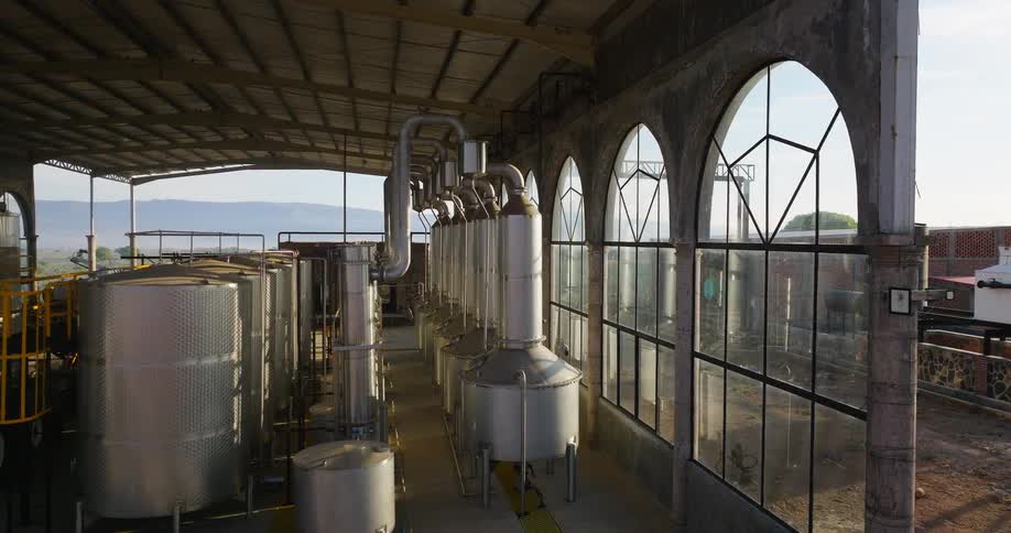 Interior of industrial distillery with stainless steel tanks connected by piping under high metal roof and arched windows overlooking mountains