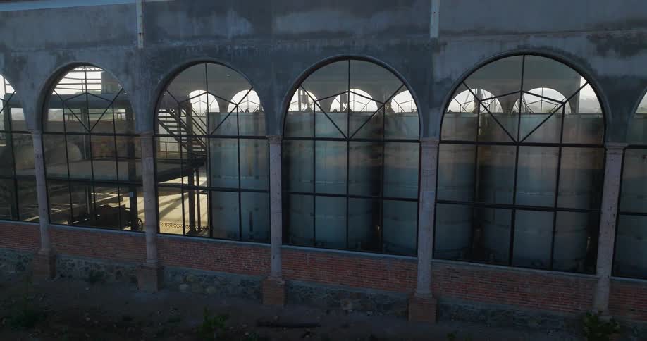 An aerial view pans slowly along the side of a concrete building with a row of tall arched windows that look into an interior containing large metal tanks before revealing an open bay where additional tanks and equipment stand