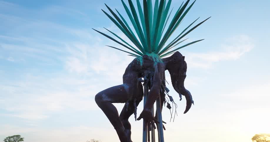 Metal sculpture depicts a human figure bent beneath agave leaves at sunset
