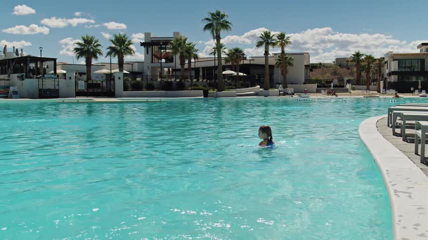 Girl swims alone in blue pool surrounded by palm trees lounge chairs and modern buildings under sunny sky
