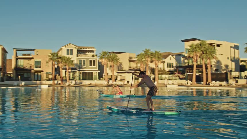 Two boys are on paddle boards moving through the water past houses