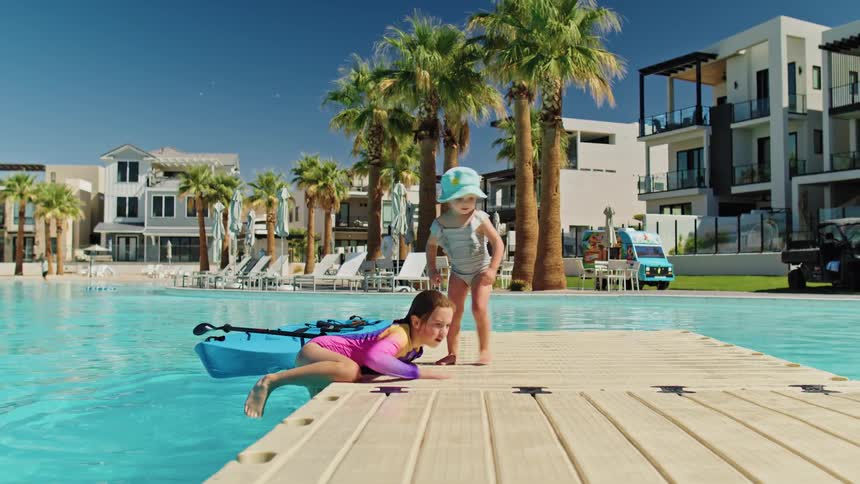 Children play on wooden dock over blue pool in sunny setting with palm trees and modern buildings in the background