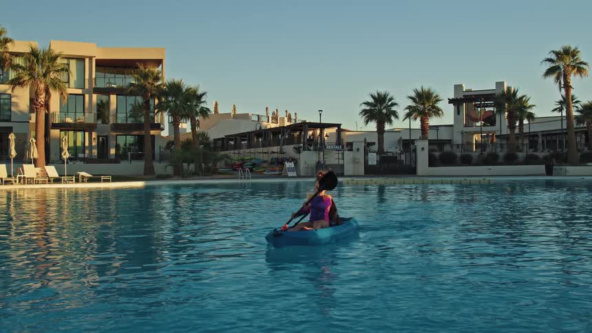 A young girl paddles a kayak across a large resort swimming pool lined with palm trees and modern buildings