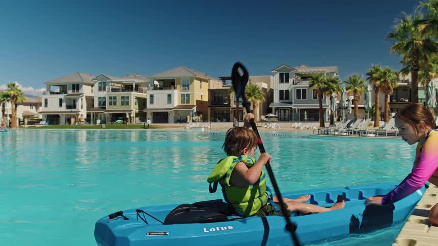 A child rides a kayak in a large public pool at midday while another girl guides and directs him through the water