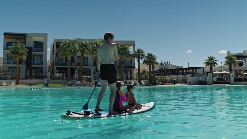 Older woman uses her smartphone to record a man and his children maneuvering a paddle board across a large pool