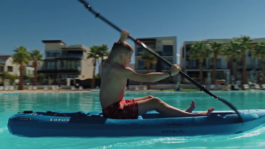 Boy paddles a blue kayak in a pool surrounded by palm trees and modern buildings