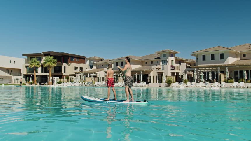Two people stand on a paddleboard in a large water lagoon one person paddles the board moves across the water and modern buildings and palm trees line the background