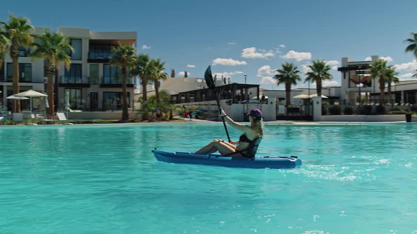 Woman paddles blue kayak on clear water surrounded by palm trees and modern buildings in sunny resort setting