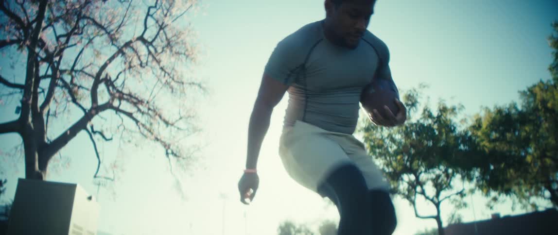A football player runs a drill in bright backlight while holding a ball on a tree lined field