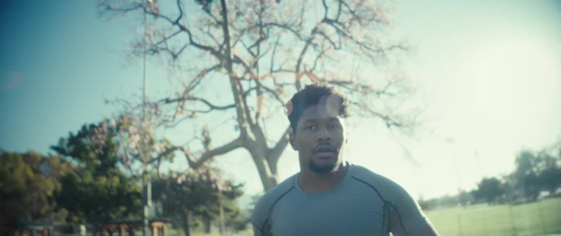 Man in gray athletic shirt looks down in outdoor setting with blue sky and leafless trees in the background