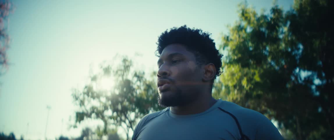 A man holding a football breathes heavily his face focused with exertion framed against the bright sky and the green leaves of park trees