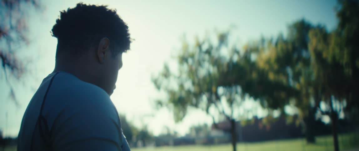 A man wearing a tshirt is seen from the shoulders up as he exercises his arms he faces away from the camera toward bright sunlight and green trees are in the background
