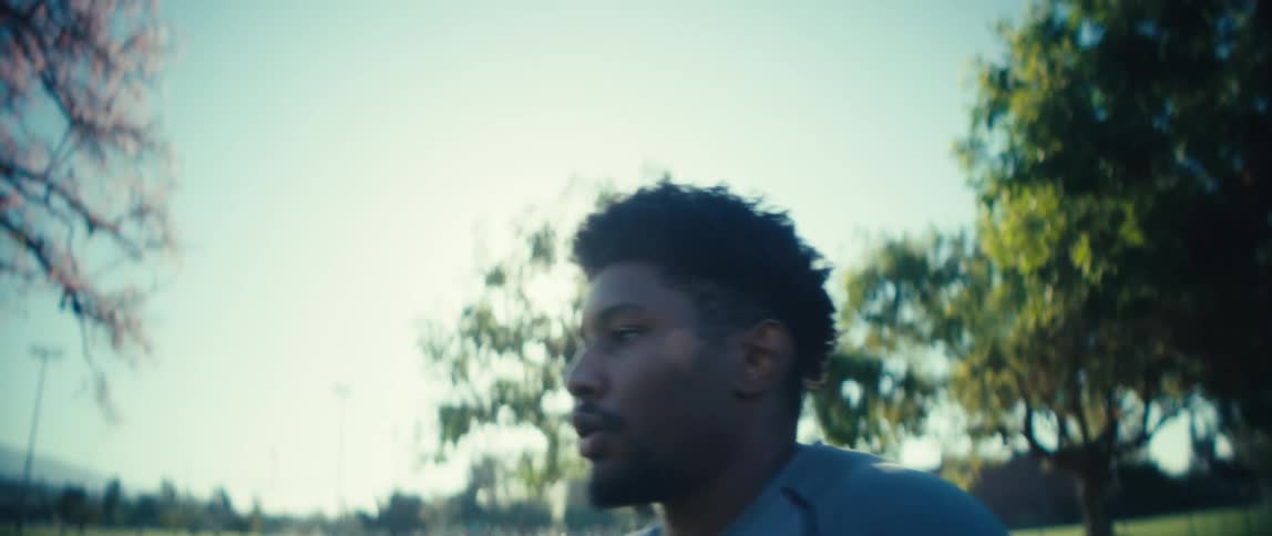 A closeup profile shot shows a man jogging or running with a focused expression in a sunny outdoor park setting he appears to be engaged in a workout breathing steadily as he moves past the camera with trees visible in the background