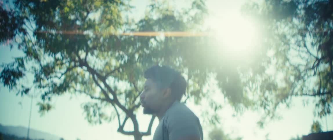 A side close view shows a man wearing a gray workout shirt running outside in a sunny park with bright lens flare shining through green tree branches overhead
