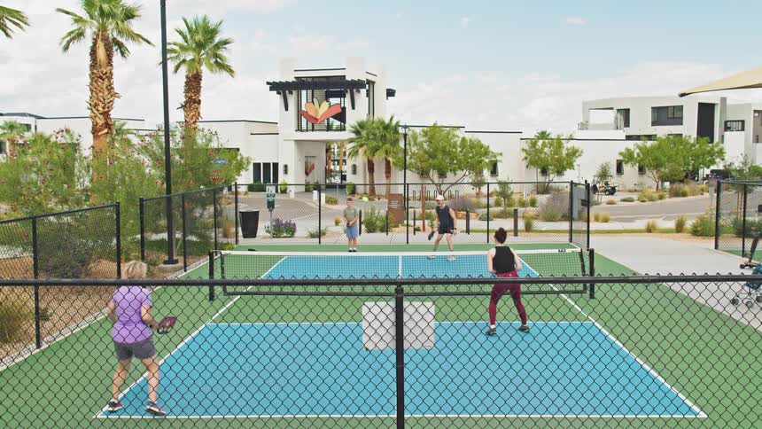 Two pairs of players engage in a pickleball game on a fenced blue and green court with palm trees and white modern resort buildings visible in the sunny background and people passing by on bicycles