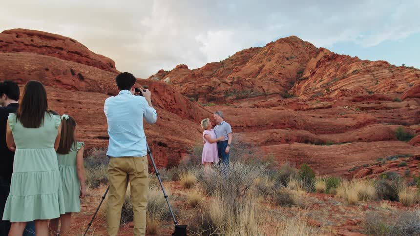 A family stands on a brush filled hillside while a man wearing a light blue shirt takes photos of a man and woman posing in front of massive layered red rock formations