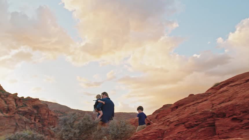Father carries a child while walking with another son on reddish rocks at sunset