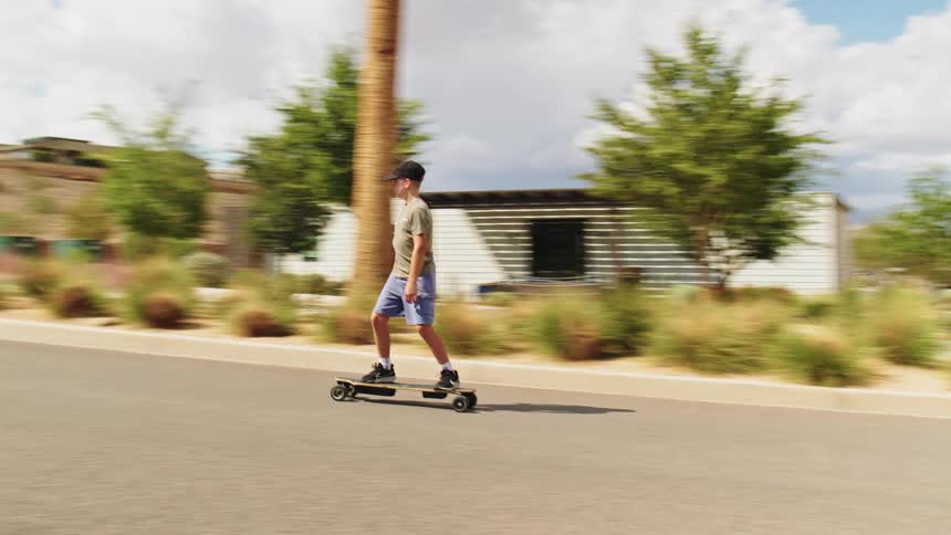 A teenage boy wearing a black baseball cap and a grey shirt rides an electric skateboard along a paved road passing palm trees and modern residential buildings in a sunny arid environment