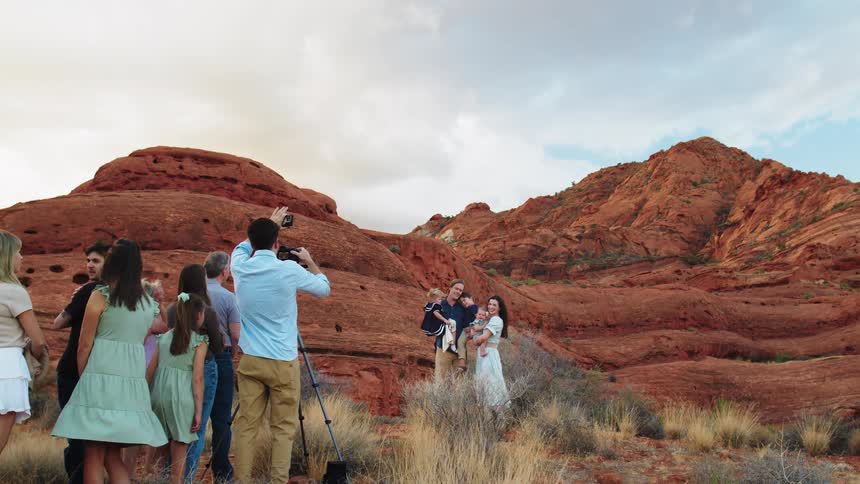 A family with the father and mother each carrying an infant stands side by side posing for a photo in a canyon while a group of people watches them at midday