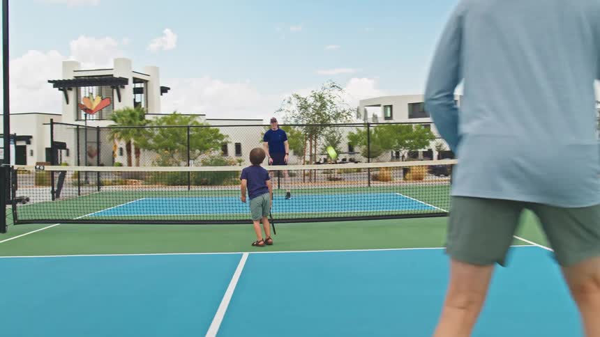 Two men and a child play pickleball on a blue and green outdoor court hitting the yellow ball over the net in a modern residential community with palm trees