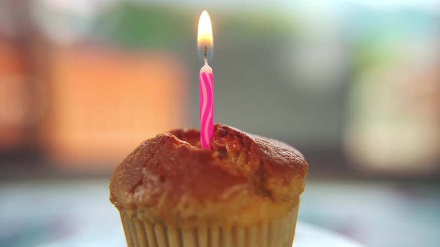 A tall pink candle inserted into the top of a muffin or cupcake is burning brightly on a blurried surface