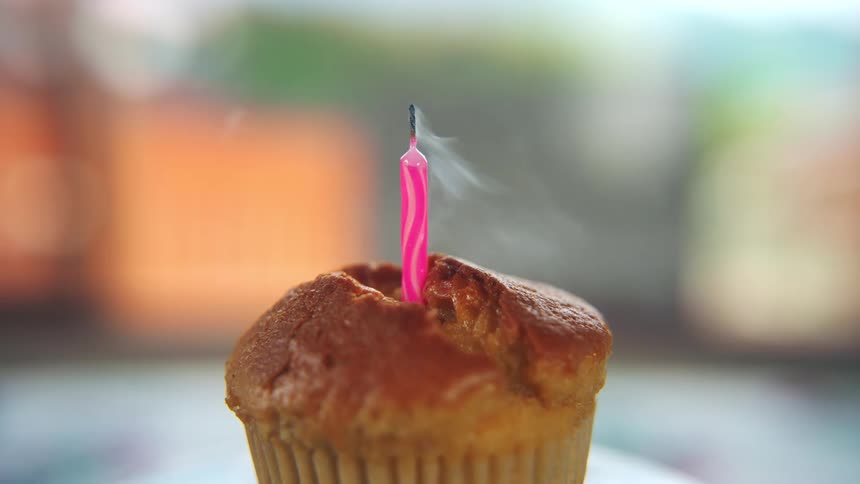 A tall pink candle inserted into the top of a muffin or cupcake is burning brightly then the flame is extinguished by a puff of air leaving a rising wisp of white smoke
