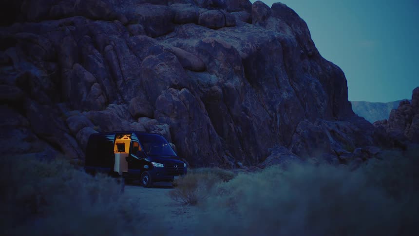 A camper van glows warmly with its door open at dusk beside dark rocky cliffs