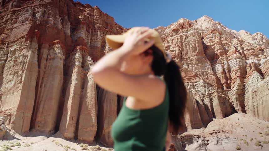 A young woman stands on a canyon wearing a baseball hat admiring the rocky formations around her under the sun