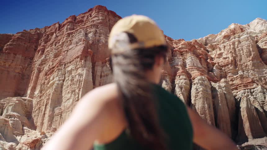 A person wearing a yellow cap and a dark tank top stands with her back to the camera looking up at a towering wall of stratified reddishorange rock formations under a bright blue sky