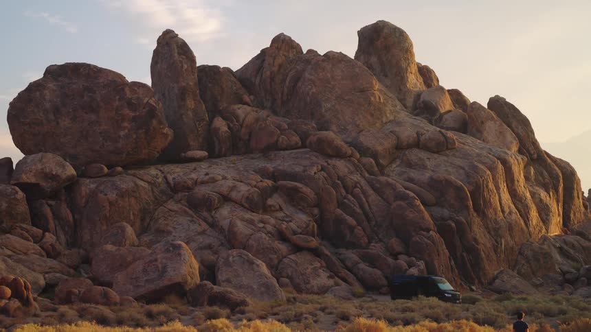 A man hikes down a mountain walking toward a van parked beside a rock in the valley at midday