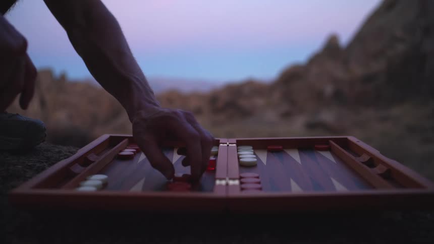 A closeup view of a backgammon board shows a persons hand picking up a checker and moving it to a new point on the wooden board the game is being played outdoors during twilight with a rocky desertlike landscape and a colorful horizon visible in the